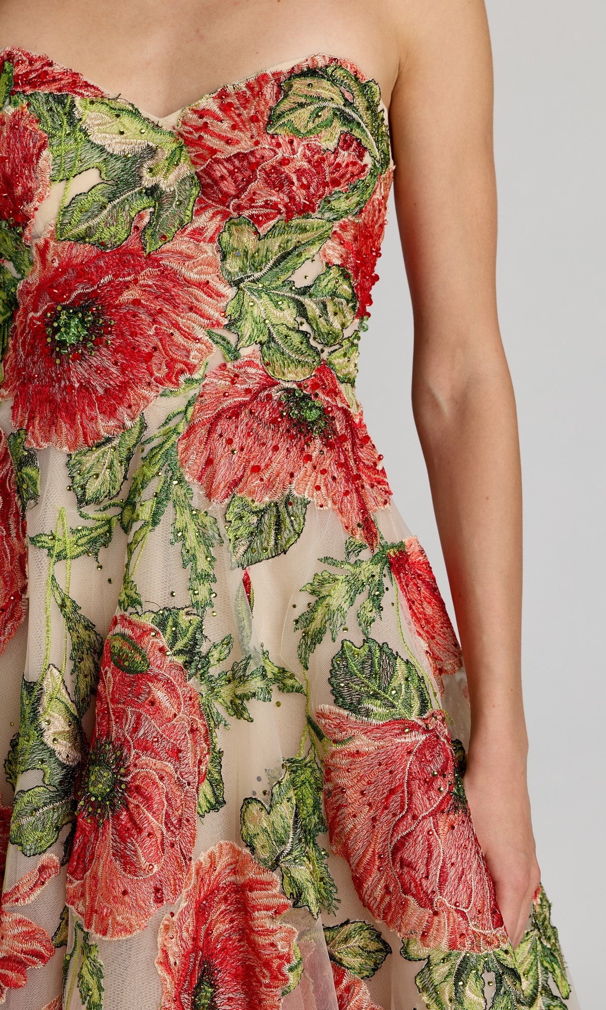 A person wearing the Mac Duggal 21300 tea-length party dress, featuring a strapless tulle design with large red and green floral embroidery, stands against a plain light background.